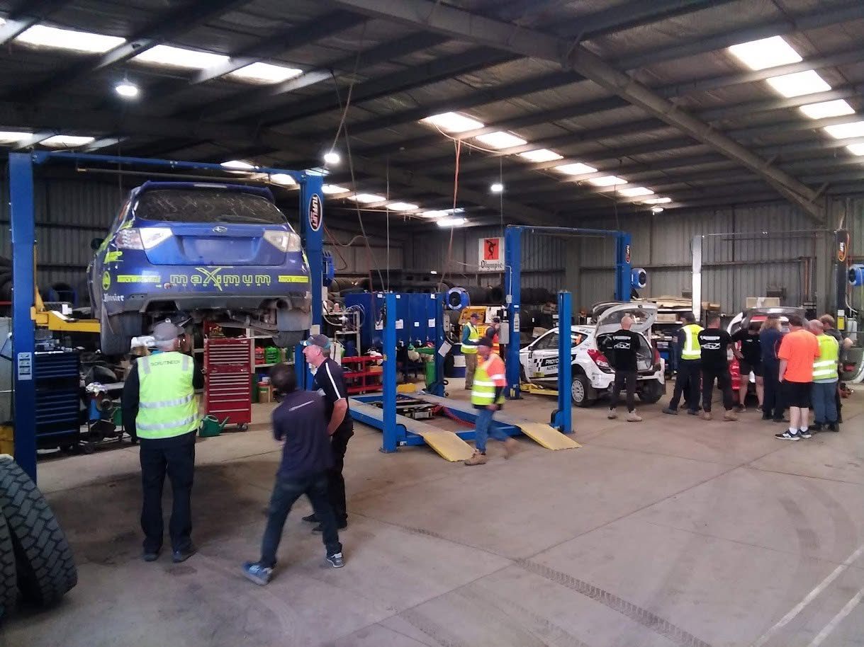 Rally cars up on the hoists being checked by scrutineers in hi-vis inside the workshop